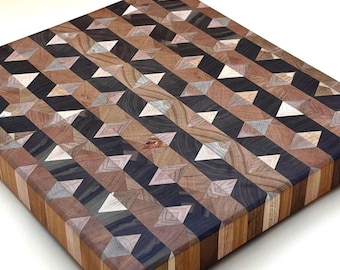 May include: A square wooden cutting board with a geometric pattern. The pattern is made up of various wood tones, including light brown, dark brown, black, and white. The board is approximately 12 inches square and has a smooth surface.