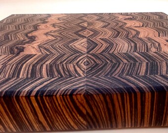 May include: A rectangular wooden cutting board with a unique geometric pattern. The board features alternating dark brown and black wood grains, creating a striking visual effect. The end-grain construction is visible, showcasing the wood's texture.