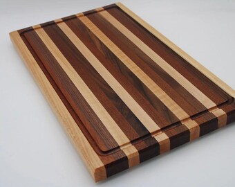 May include: Rectangular wooden cutting board with a striped pattern of light and dark brown wood. The board has a juice groove around the edge and is made of various wood types. A kitchen item.