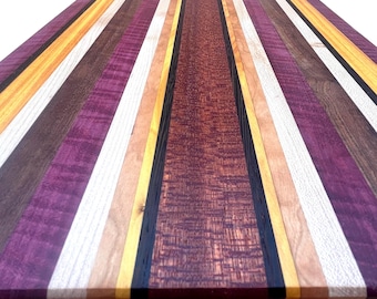 May include: A rectangular wooden cutting board with a striped pattern. The stripes alternate between shades of purple, brown, yellow, and natural wood tones. The board has a smooth, polished finish, showcasing the natural wood grain. The edges are also finished with a contrasting wood border.