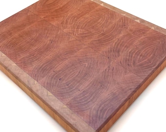 May include: A square, end-grain cutting board with a rich, reddish-brown hue. The board features a border of lighter wood and measures approximately 12 inches by 12 inches. The wood grain pattern is visible throughout the surface.