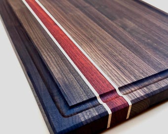 May include: A rectangular wooden cutting board with a dark wood grain. A red stripe with white accents runs down the center. The board has a juice groove and is made of multiple wood types.