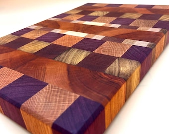 May include: A rectangular wooden cutting board with a checkerboard pattern. The board features a variety of wood tones, including reddish-brown, purple, and light brown. The end-grain construction is visible, showcasing the wood's texture and grain.