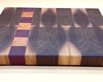 May include: A rectangular wooden cutting board with a unique end-grain design. The board features a combination of dark and light wood tones, with a central strip of purple and orange blocks. The overall pattern creates a visually appealing and functional kitchen accessory.