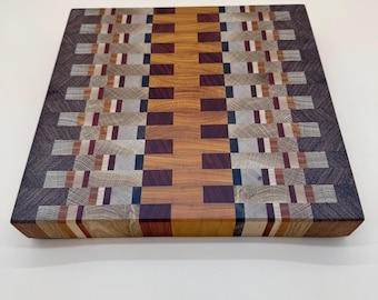 May include: A rectangular end-grain cutting board with a mosaic pattern. The board features a central section of light brown wood, flanked by sections of darker wood, including shades of brown, gray, and red. The board is approximately 12 inches square.
