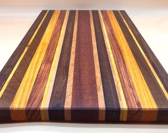 May include: A rectangular wooden cutting board with a striped pattern. The stripes alternate between dark brown, light brown, yellow, and tan. The board is made of various types of wood and has a smooth finish.
