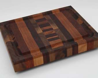 May include: A rectangular wooden cutting board with a juice groove. The board is made of various wood tones, including light brown, dark brown, and reddish-brown. The wood grain is visible, and the board has a smooth finish. The cutting board is a kitchen item.
