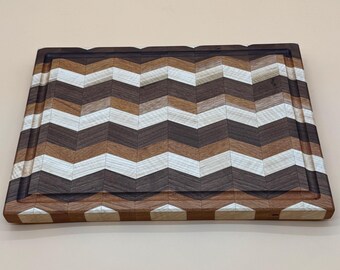 May include: A rectangular wooden cutting board with a chevron pattern. The board features alternating stripes of light and dark wood tones, including white, brown, and walnut. A dark brown border surrounds the cutting surface, with a juice groove.