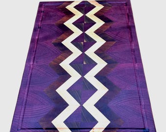 May include: A rectangular wooden cutting board with a purple border and a zig-zag pattern in the center. The zig-zag design is made of light wood and dark wood. The board has a juice groove around the edge.