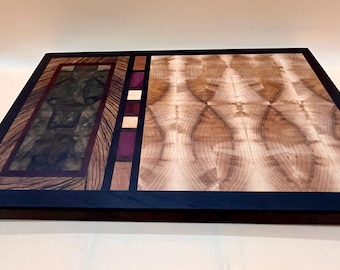 May include: A rectangular wooden cutting board with a dark blue border. The board features a section of dark green and brown wood, and a section of light brown wood with a unique pattern. Accents of purple and cream-colored wood are also present.