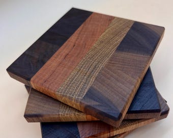 May include: A stack of four square wooden coasters featuring a striped pattern of alternating dark walnut, reddish-brown cherry, and textured light oak wood segments. Each coaster has a smooth, flat surface with clean, straight edges and a polished finish.