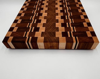 May include: A rectangular wooden cutting board with a geometric pattern. The board features alternating squares and rectangles of light and dark wood, creating a visually appealing design. The edges are angled, and the board is set against a white background.