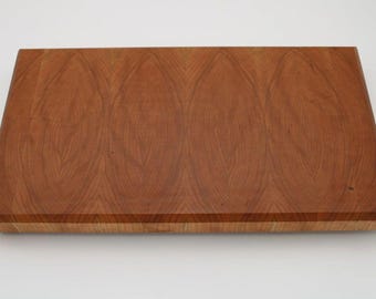 May include: A rectangular wooden cutting board with a rich, reddish-brown color. The wood grain pattern is prominent, creating a visually appealing surface. The board has a beveled edge and is likely used for food preparation.