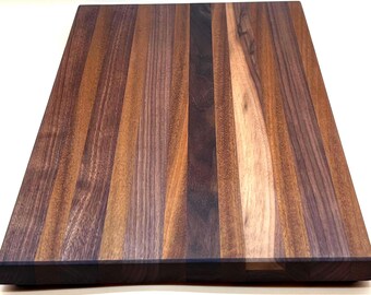 May include: A rectangular wooden cutting board with a striped pattern. The board is made of various shades of brown wood, including dark walnut and lighter tones. The wood grain is visible, and the board has a smooth, finished surface.