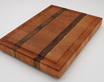 May include: A rectangular wooden cutting board with a light brown and dark brown striped design. The board has a recessed juice groove around the perimeter. The wood grain is visible, and the board appears to be made of cherry wood.