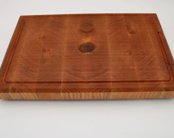 May include: A rectangular wooden cutting board with a reddish-brown hue. The board features a juice groove around the perimeter and is constructed from end-grain wood. The wood grain is visible throughout the board.