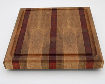 May include: Square wooden cutting board with a juice groove. The board features alternating light and dark wood grain patterns, with a prominent dark red stripe down the center. The edges are also made of alternating wood colors.
