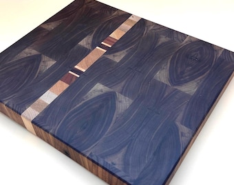 May include: A square, end-grain cutting board with a dark blue and brown wood pattern. A diagonal strip of lighter wood with various colors crosses the board. The board is made of multiple wood pieces, creating a unique design.