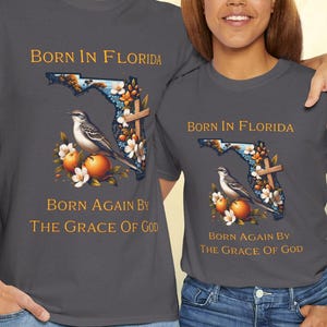 May include: Two charcoal gray t-shirts with the text "BORN IN FLORIDA" and "BORN AGAIN BY THE GRACE OF GOD". The shirts feature a Florida outline with a bird, flowers, and oranges. The people are wearing blue jeans.