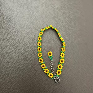 May include: A beaded bracelet featuring a sunflower design. The bracelet is made of yellow, brown, and green beads, with a silver-colored clasp. The sunflower beads are arranged in a repeating pattern around the wrist. The bracelet is approximately 7 inches long.