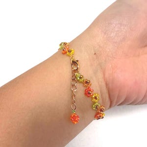 May include: A beaded bracelet featuring small, colorful flower-shaped beads in shades of yellow, orange, and green. The bracelet has a gold-colored clasp and an adjustable chain. The bracelet is approximately 7 inches (17.78 cm) long.