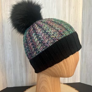May include: A knitted beanie with a black faux fur pom-pom. The hat features a black ribbed brim and a colorful patterned crown with shades of purple, green, and blue. The hat is displayed on a wooden head form.