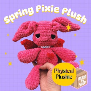 May include: A handmade pink crochet plushie in the shape of a pixie bunny with large ears and red wings. The plushie has a heart-shaped detail and a beaded necklace that spells out a word. The text "Spring Pixie Plush" is at the top.