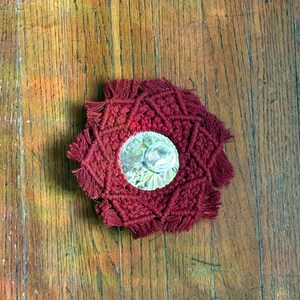May include: A burgundy macrame coaster with a circular mirror center. The coaster has a geometric pattern and fringe edges. The coaster is on a wooden surface.