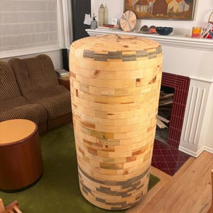 Peut inclure: Une table d'appoint cylindrique en bois, construite à partir de blocs rectangulaires superposés de bois clair. La table présente un aspect de grain de bois naturel avec des accents de gris et de vert. Elle est posée sur un tapis vert.