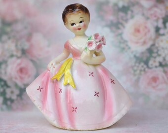 Może przedstawiać: Vintage porcelanowa figurka młodej dziewczyny w różowej sukience trzymającej bukiet różowych kwiatów. Sukienka ma żółtą szarfę i jest ozdobiona małymi czerwonymi krzyżykami. Figurka ma brązowe włosy i jest osadzona na rozmytym kwiatowym tle.