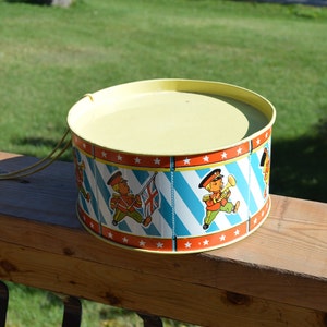 May include: A yellow toy drum with a blue and white striped design and red stars. The drum has a rope handle and features a marching band scene with three figures.