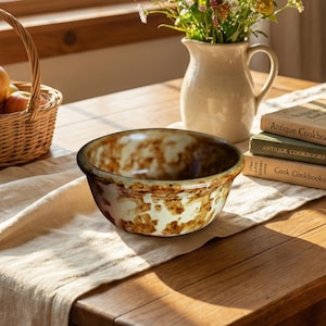 May include: A ceramic mixing bowl with a brown and cream mottled pattern sits on a beige linen tablecloth. A wicker basket of apples, a cream-coloured jug with flowers, and antique cookbooks are also on the wooden table.