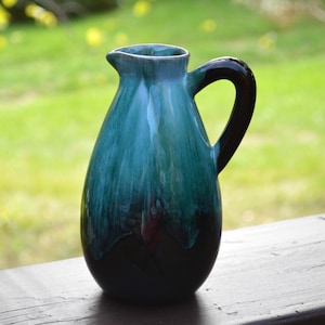 May include: A ceramic jug with a dark handle and a spout. The jug has a glossy finish with a gradient of colours, transitioning from a dark base to teal and green hues. The jug is likely used for serving beverages.