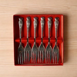 May include: A set of six silver-toned metal forks arranged in a red rectangular presentation box. Each fork features a decorative handle with an embossed thistle design at the top and three tines at the base, all neatly held in place.