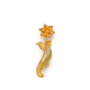 May include: A gold-tone brooch featuring a yellow flower with green leaves. The flower is made of rhinestones and the leaves are accented with smaller rhinestones.