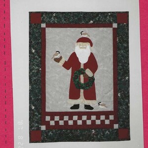 May include: A red and white quilted wall hanging featuring a Santa Claus figure holding a wreath and surrounded by birds. The background is a green and white checkered pattern.