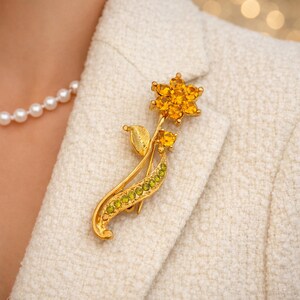 May include: A gold-toned floral brooch with a flower and leaves design. The flower features yellow gemstones, while the stem has green gemstones. The brooch is pinned to a cream-colored blazer.