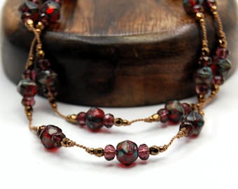 Op de afbeelding: Een ketting met gefacetteerde rode en donkergekleurde kralen afgewisseld met kleinere metallic bronzen kralen. De kralen zijn geregen op een dunne, gedraaide bronskleurige draad, wat zorgt voor een verfijnde uitstraling met een herhalend patroon.