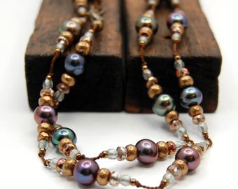 Peut inclure: Un collier multi-brins composé de perles irisées violettes et bleues foncées, alternées avec des perles métalliques bronze texturées et de petites perles en verre transparent, le tout enfilé sur un fin cordon marron.