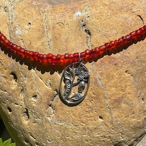 May include: A red beaded necklace with a silver pendant. The pendant features a butterfly and plant design within an oval frame. The necklace is displayed on a textured, light brown rock.
