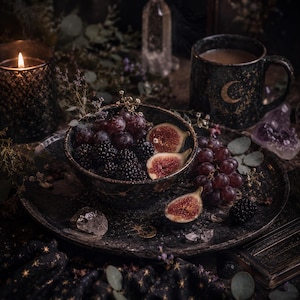 May include: A black ceramic bowl and matching plate decorated with gold stars and crescent moons, filled with fresh blackberries, red grapes, and sliced figs. A matching mug, a lit candle in a glass holder, and clear crystals complete the set.