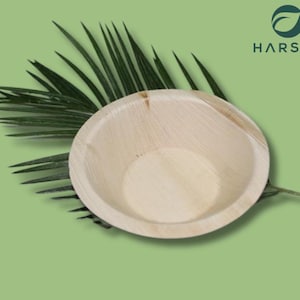 May include: A round, light-colored bowl made from natural materials, possibly palm leaf, sits on a bed of green palm fronds. The bowl has a smooth interior and a slightly textured exterior. The background is a solid, light green color. The logo "HARSHU" is in the upper right corner.