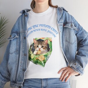 Puede incluir: Camiseta blanca con una ilustración de acuarela de un gato dentro de una bolsa de patatas fritas, comiendo una patata frita. El texto "ARE LIKE POTATO CHIPS YOU CAN NEVER HAVE JUST ONE" está impreso en azul alrededor de la imagen.
