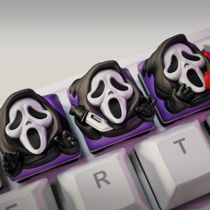Op de afbeelding: Close-up van een mechanisch toetsenbord met aangepaste keycaps met het Ghostface-masker uit de Scream-franchise. De keycaps zijn zwart, wit en paars, met het maskerontwerp. De toetsen van het toetsenbord zijn wit.