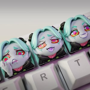May include: Three custom keycaps featuring a stylized anime character with teal hair and expressive faces. The keycaps are on a white keyboard, with the "nom nom figures" logo visible in the corner. Each keycap has a different facial expression.