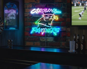 May include: A neon sign displaying the Carolina Panthers logo and team name in vibrant colors against a brick wall. The sign is illuminated with blue, green, and pink lights. A framed jersey and a dartboard are visible.