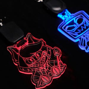 May include: Two illuminated keychains with cartoon character designs. One keychain glows red, featuring a character with a hat and staff. The other glows blue, showcasing a robot-like character. Both are on black lanyards.