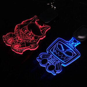 May include: Two glowing acrylic keychains with cartoon character designs. One keychain glows red, the other glows blue. Both have a black leather strap and metal keyring. The keychains are on a black background.