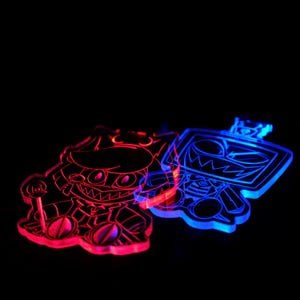 May include: Two acrylic keychains with cartoon character designs. One keychain glows red, the other glows blue. The keychains have a dark background and are illuminated with neon-like light.