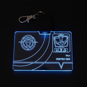 Puede incluir: Un llavero rectangular azul brillante con un diseño de personaje de dibujos animados. El llavero tiene un cierre plateado y un soporte negro. El diseño incluye un logotipo y el texto "Vox VOXTEK CEO".